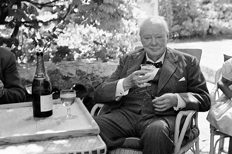 Winston Churchill seated outdoors with a cigar in his left hand and a Champagne glass beside an open bottle.